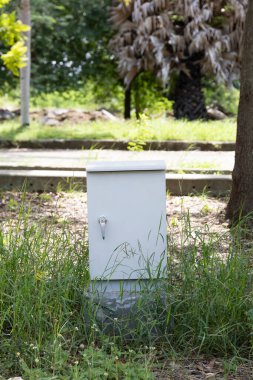 Beyaz elektrik kutusu çimlerin ve yeşilliklerin arasında duruyor. Doğayı altyapıyla harmanlayarak tesise güvenli bir sistem sağlıyor. Güvenliği ve güvenilir hizmetleri sağlıyor..