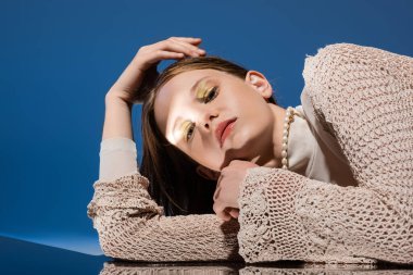 Portrait of woman in knitted cardigan looking at camera near reflective surface on gradient blue background  clipart