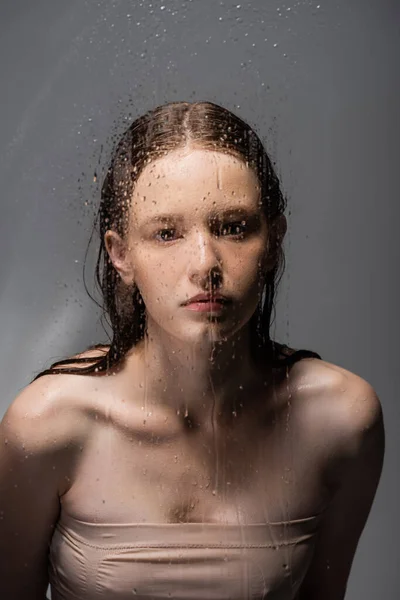 Young woman looking at camera behind wet glass on grey background