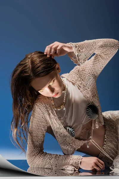 Fair haired model in knitted cardigan and pearl necklace looking at reflective surface on blue background