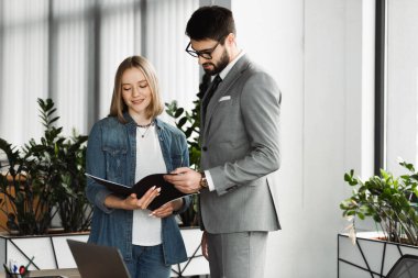 Smiling woman holding paper folder with resume near businessman in formal wear during job interview in office  clipart