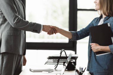 Cropped view of smiling woman with resume shaking hand of businessman during job interview in office clipart