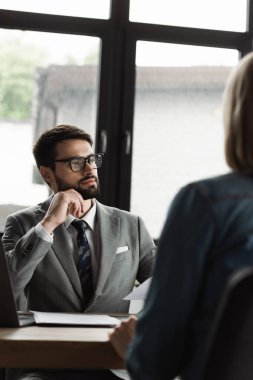 Manager in formal wear sitting near papers and blurred candidate on job interview  clipart