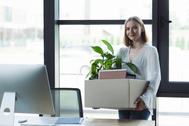Smiling intern holding carton box and looking at camera near computer in office  clipart