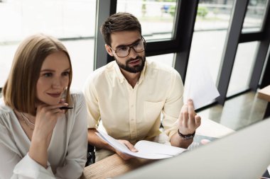 Businessman holding papers near computer and blurred intern in office  clipart