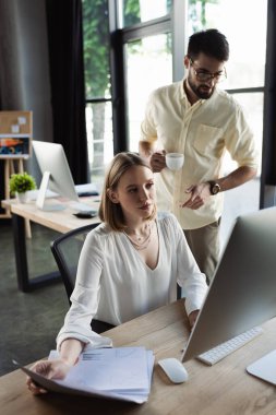 Intern looking at computer near papers and businessman with coffee in office  clipart