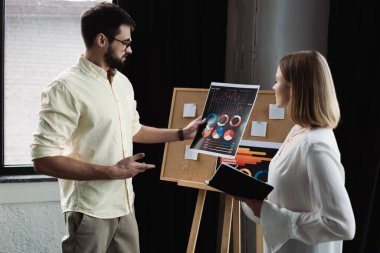 Side view of businessman pointing at charts near intern with notebook in office  clipart