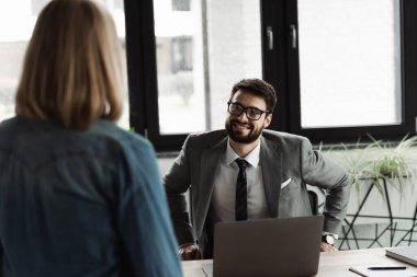 Smiling businessman in eyeglasses looking at blurred job seeker in office  clipart