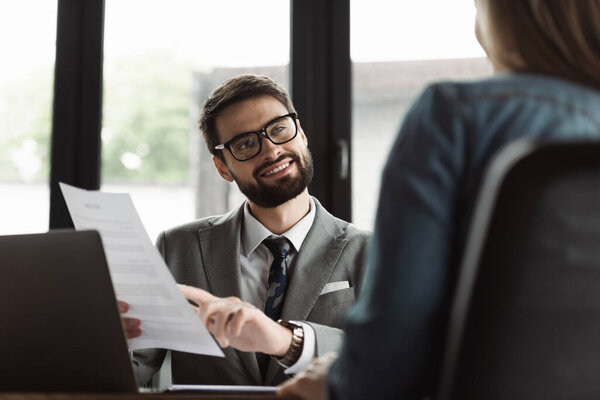Smiling manager pointing at resume near blurred candidate and laptop in office 