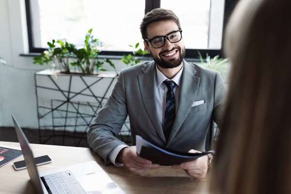 Cheerful businessman holding paper folder with resume near blurred candidate and devices in office 