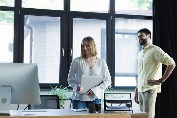Smiling woman holding laptop near working table and businessman during internship in office 