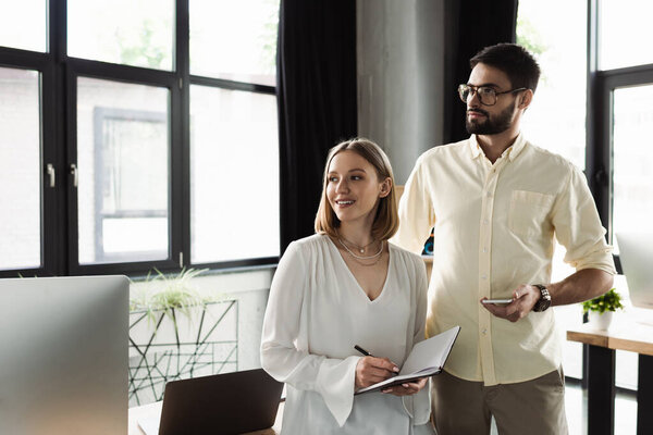 Businessman holding smartphone near smiling intern writing on notebook in office 