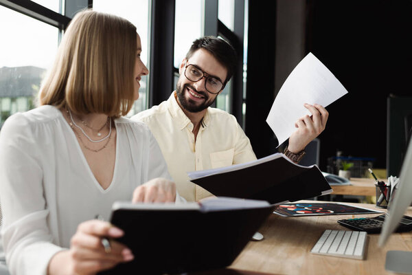 Positive manager holding paper folder and looking at new worker with notebook in office 