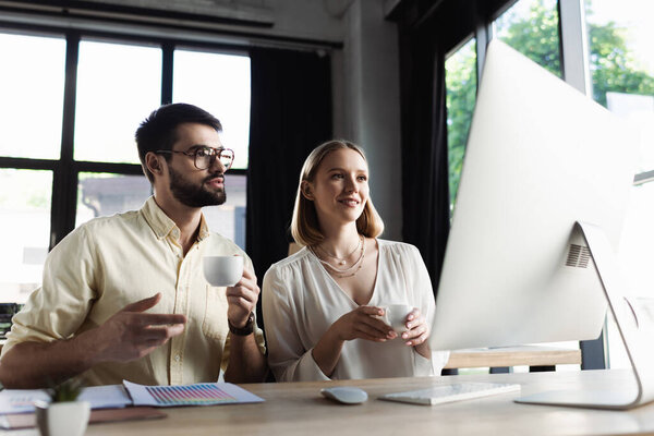 Businessman holding coffee and talking to intern near computer in office 