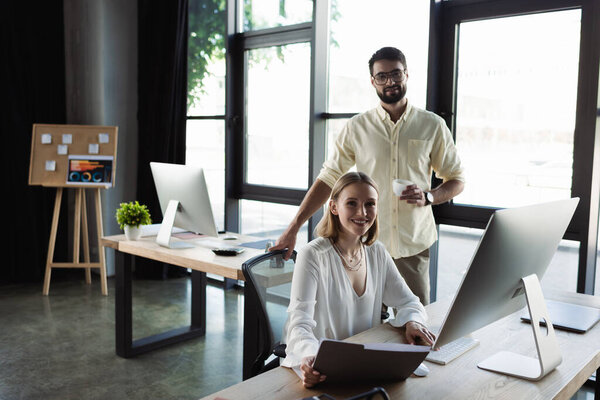 Smiling intern and businessman with coffee looking at camera near computer in office 
