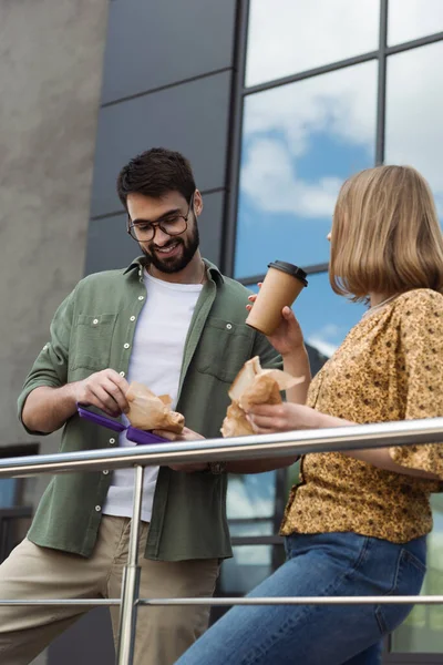 笑顔のビジネスマンがコーヒーと同僚の近くのサンドイッチとランチボックスを保持する都市部の通りに行く — ストック写真