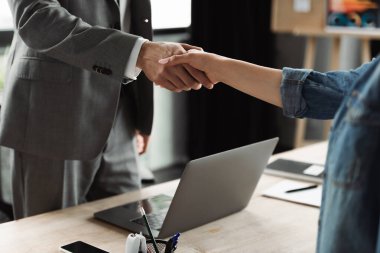 Cropped view of job seeker and businessman shaking hands near devices in office  clipart