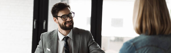Positive businessman in formal wear looking at blurred woman during job interview in office, banner 