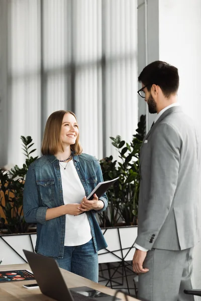 Smiling job seeker holding paper folder near businessman during interview in office