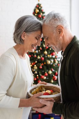 Gülümseyen orta yaşlı çift ellerinde hasır sepetle süslenmiş Noel ağacının yanında