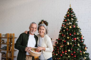 smiling middle aged couple holding wicker basket with baubles near decorated christmas tree clipart