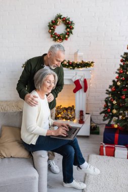 Orta yaşlı bir adam, yılbaşı ağacının yanında dizüstü bilgisayar kullanarak neşeli karısına sarılıyor. 