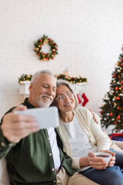 Orta yaşlı bir adam evde mutlu bir eşle selfie çekiyor.