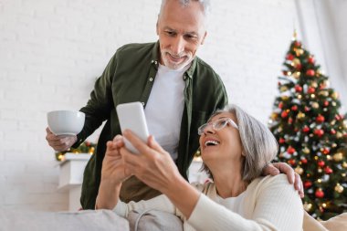 Olgun bir adam elinde çay fincanıyla karısının yanındaki akıllı telefona bakıyor ve bulanık Noel ağacına bakıyor.