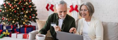 Mutlu, olgun bir adam, Noel 'de online alışveriş yaparken karısının yanında dizüstü bilgisayarla kredi kartı tutuyor.
