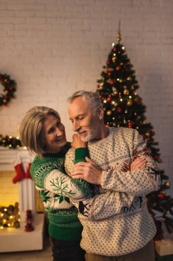 cheerful woman in festive sweater hugging pleased mature husband near decorated christmas tree in evening clipart