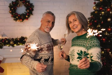 Neşeli orta yaşlı çift Noel arifesinde ellerinde parlak maytaplar ve şampanya kadehleriyle