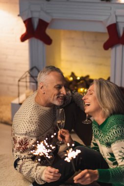 Mutlu orta yaşlı bir adam elinde şampanya kadehiyle ve neşeli karısına bakıyor Noel arifesinde parlak maytapların yanında.