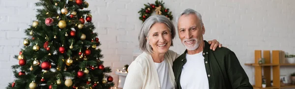 幸福的中年夫妇在装饰过的圣诞树旁微笑 高举旗帜 — 图库照片