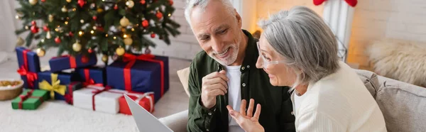 stock image smiling middle aged woman pointing at happy husband and having video call on laptop during christmas, banner