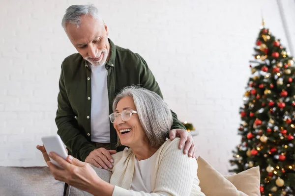 stock image happy mature man looking at smartphone near cheerful wife and blurred christmas tree