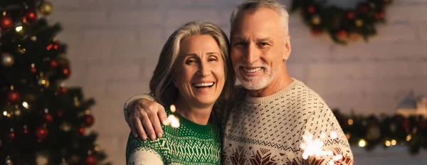 幸せな成熟したカップルで休日のセーターはクリスマスの前夜に輝く輝きの近くで笑っていますバナー — ストック写真
