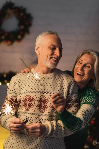 Positive Middle Aged Woman Hugging Bearded Husband While Holding Sparkler — Stock Photo, Image