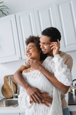 Mutlu Afro-Amerikan erkeği, mutfakta beyaz önlüklü kazak giyen heyecanlı kız arkadaşını kucaklıyor.