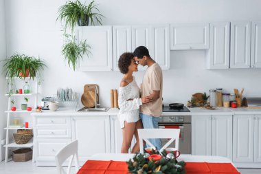 Beyaz mobilyalarla modern mutfakta kucaklaşan seksi Afrikalı Amerikalı çiftin yan görünüşü