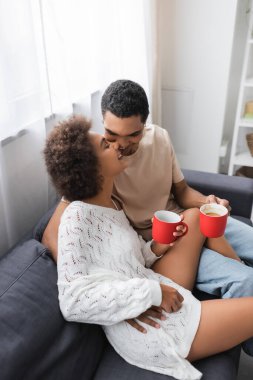 Kırmızı bardaklı Afro-Amerikan çiftin evdeki kanepede gülümsediği yüksek açı.