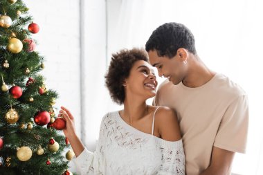 Mutlu Afro-Amerikalı kadın evde Noel ağacı süslerken genç erkek arkadaşına bakıyor.