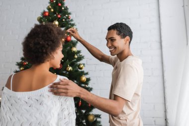 Neşeli Afrikalı Amerikalı adam beyaz önlüklü kazak ve Noel ağacı süsleme kız arkadaşına dokunuyor.