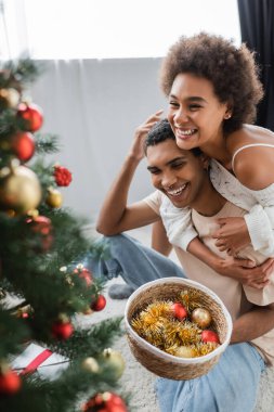 cheerful african american woman embracing boyfriend holding baubles near blurred christmas tree clipart