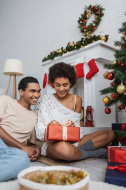 happy and sexy african american woman sitting on floor with christmas present near fireplace and smiling boyfriend  clipart