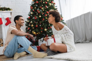 Neşeli Afrikalı Amerikalı çift Noel ağacının yanında oturmuş birbirlerine bakıyorlar. 