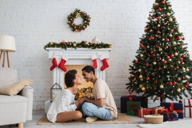 Afro-Amerikalı mutlu çiftin yan görüntüsü yerde, süslü şömine ve noel ağacının yanında oturuyor.