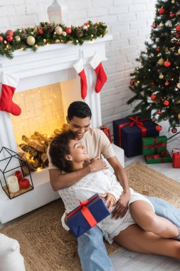 Afrikalı Amerikalı erkek kız arkadaşına sarılıyor Noel hediyesiyle birlikte süslü şöminenin yanında oturuyor.