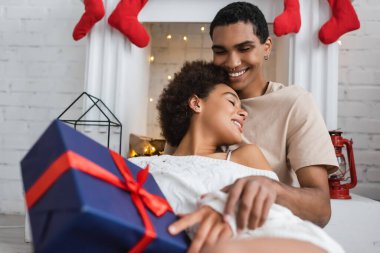 sexy woman holding christmas present near smiling african american boyfriend sitting at fireplace clipart