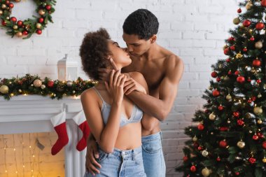 Genç ve seksi Afrikalı Amerikalı çift Noel ağacı ve süslü şöminenin yanında öpüşüyor.