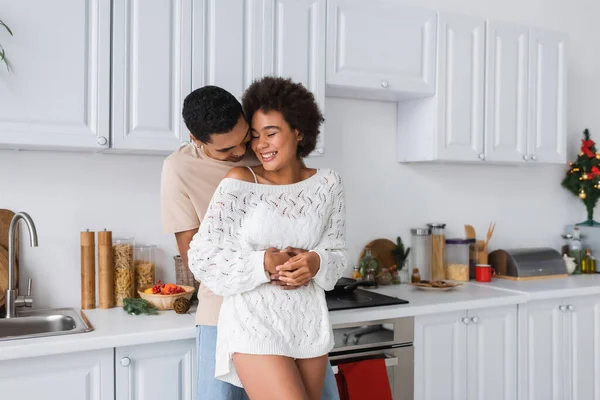 Αφροαμερικάνος Άντρας Αγκαλιάζει Χαρούμενη Φιλενάδα Λευκό Πλεκτό Πουλόβερ Στην Κουζίνα — Φωτογραφία Αρχείου